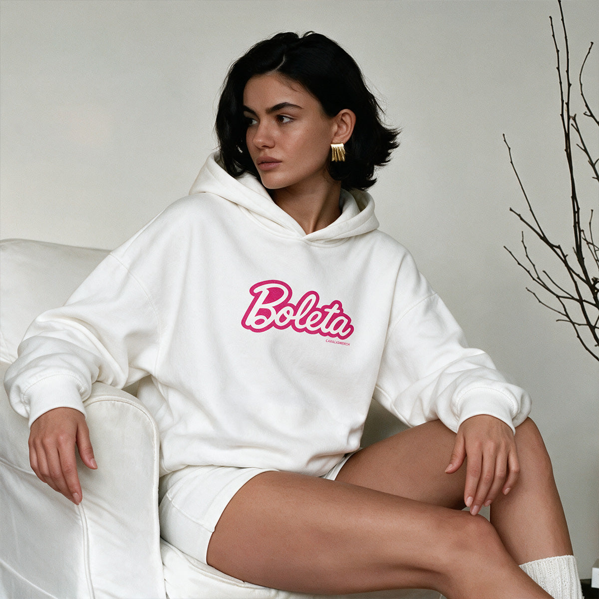 Mujer sentada llevando la sudadera con capucha blanca «Boleta» de Caracas Merch con el logotipo rosa